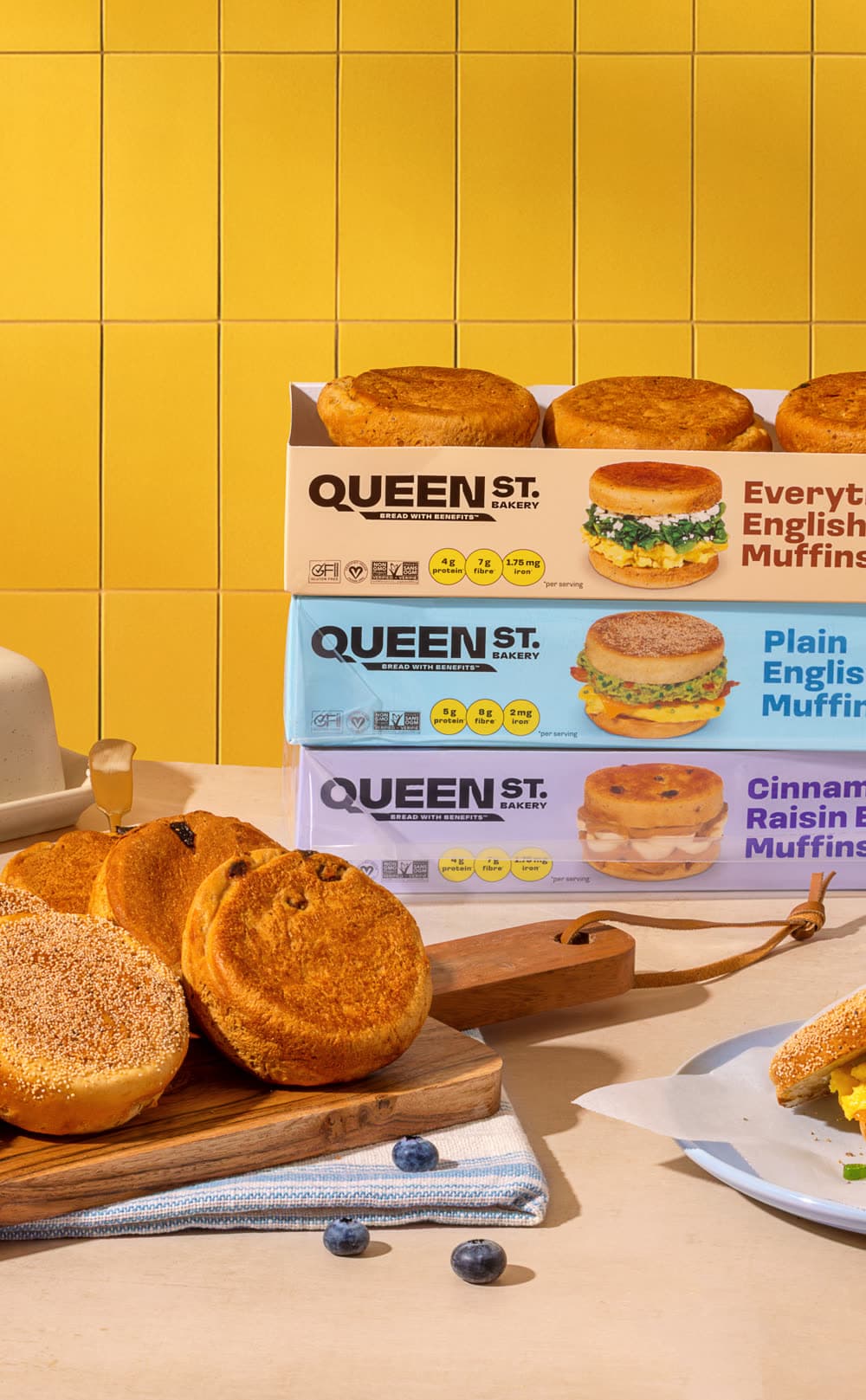 Stacked Queen St. Bakery English muffin boxes and fresh bagels on a wooden board, with a breakfast sandwich in the foreground.